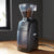 This is Encore electric grinder from Baratza. The image displays a black electric grinder laid in a wooden table behind black walls.